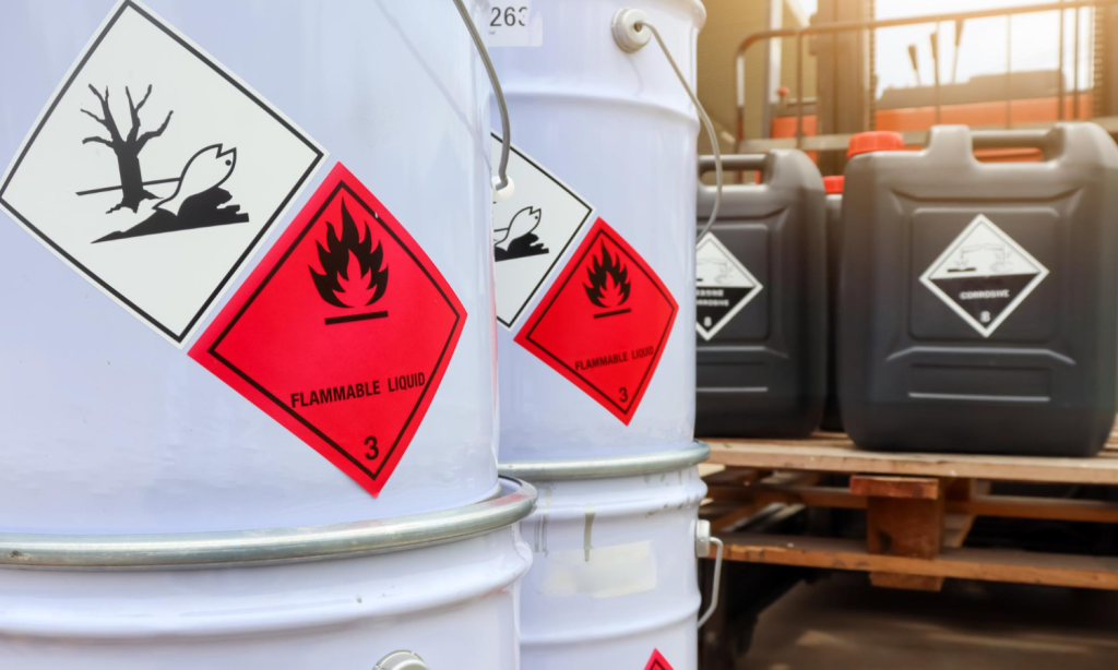 Tambores e embalagens de produtos químicos identificados com pictogramas GHS de inflamável, corrosivo e perigo ao meio ambiente, reforçando a importância da rotulagem e da conformidade com normas de segurança.
